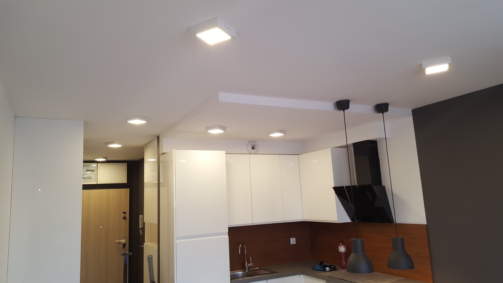 Nowoczesna kuchnia z białymi, lakierowanymi szafkami, czarnym okapem i trzema wiszącymi lampami z szarymi kloszami, oświetlona kwadratowymi lampami sufitowymi LED.