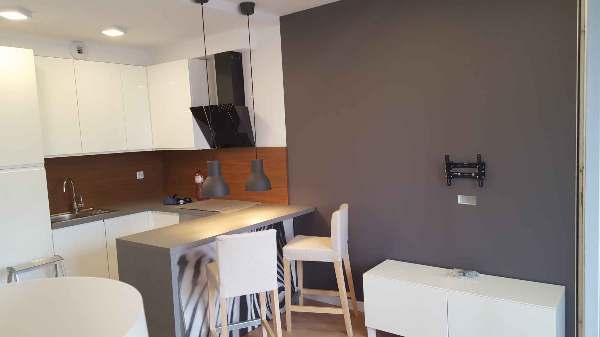 Nowoczesna kuchnia z wyspą i hokerami w Krakowie, z zamontowanym uchwytem na telewizor na szarej ścianie.