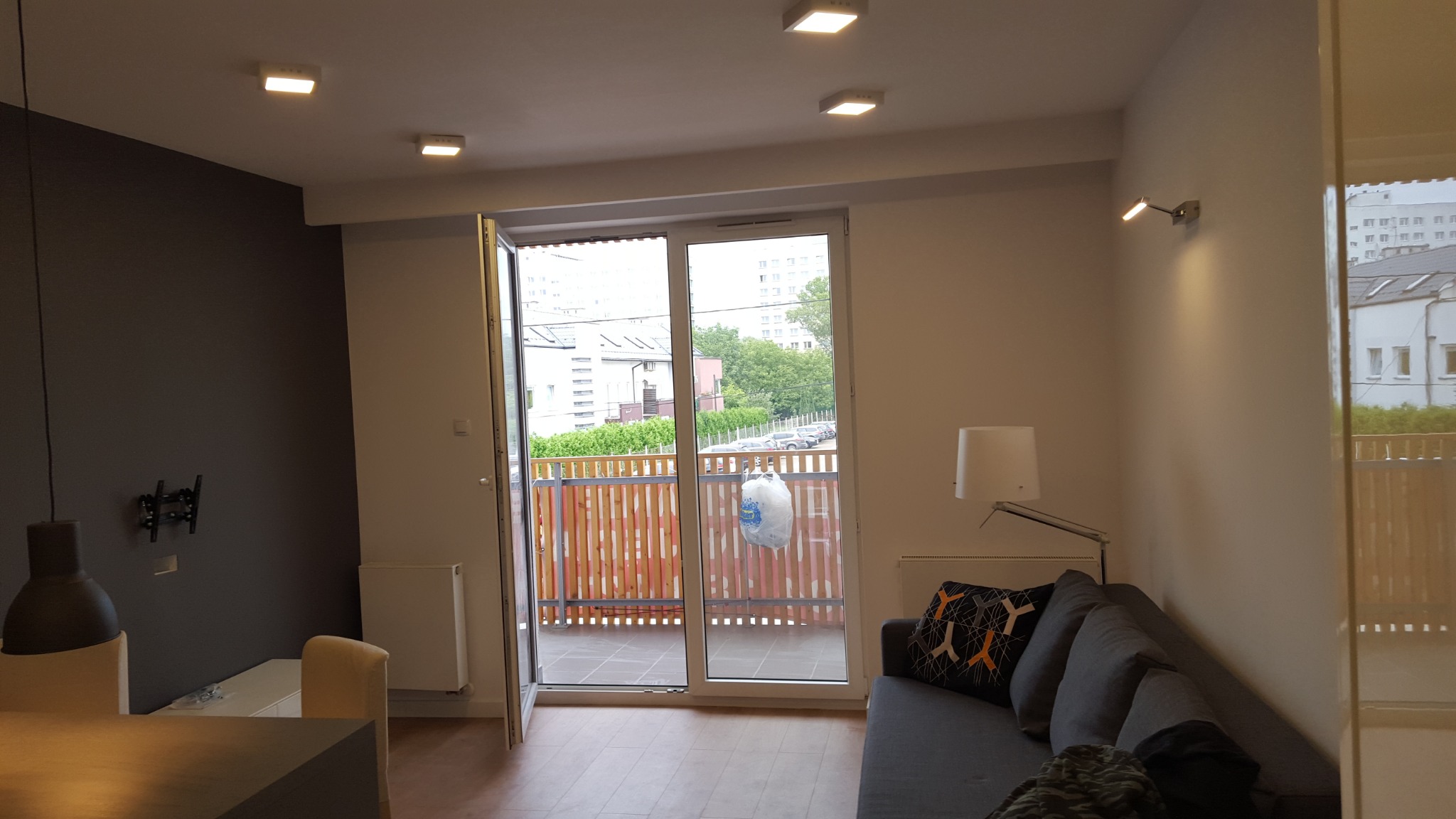 Jasne wnętrze mieszkania z otwartym balkonem, widoczny szary narożnik, lampa podłogowa i uchwyt na telewizor na ciemnej ścianie.