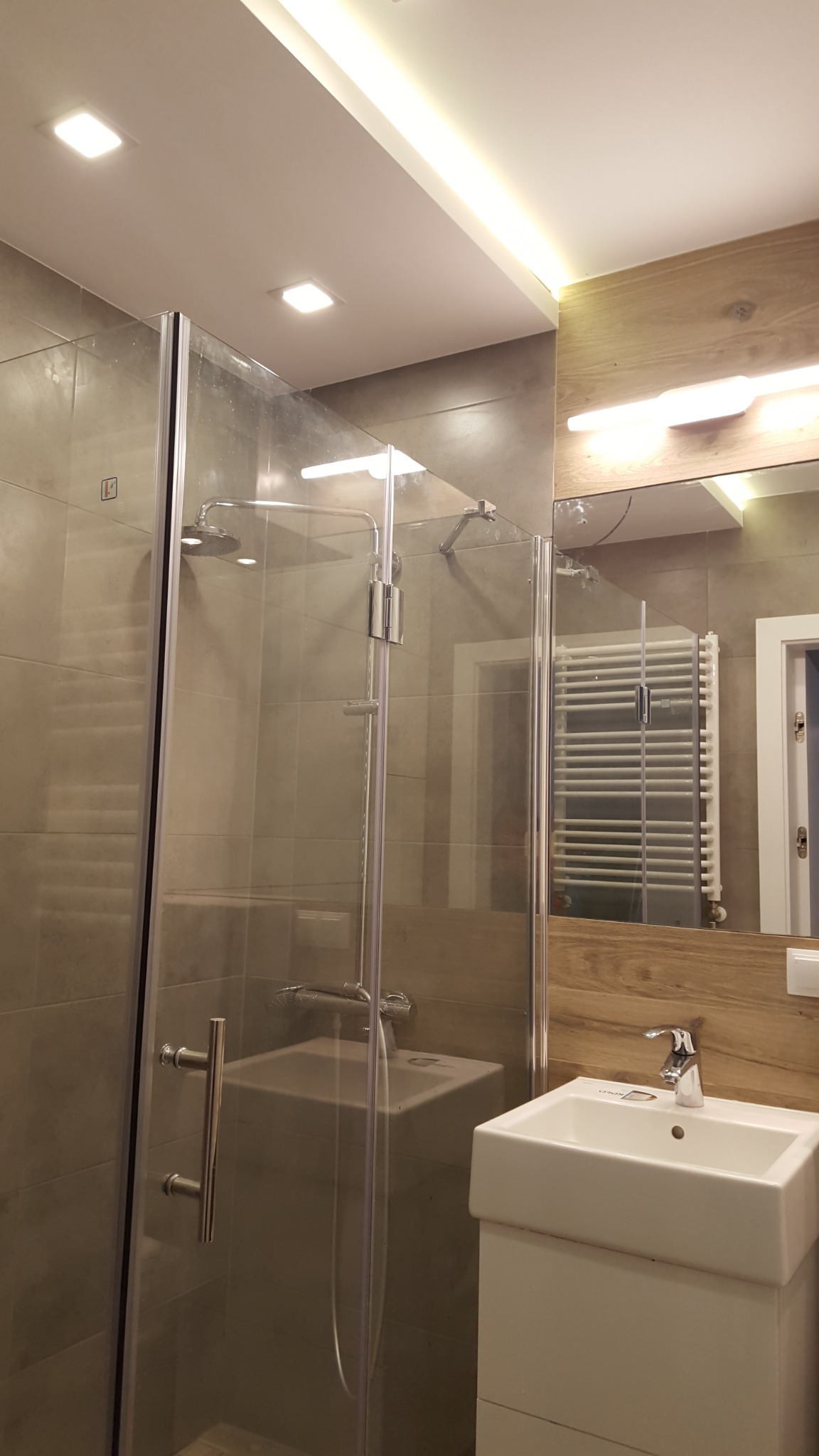 Nowoczesna łazienka po remoncie: kabina prysznicowa ze szklanymi drzwiami, umywalka z szafką i lustro z drewnopodobną okładziną, oświetlenie punktowe i ledowe.