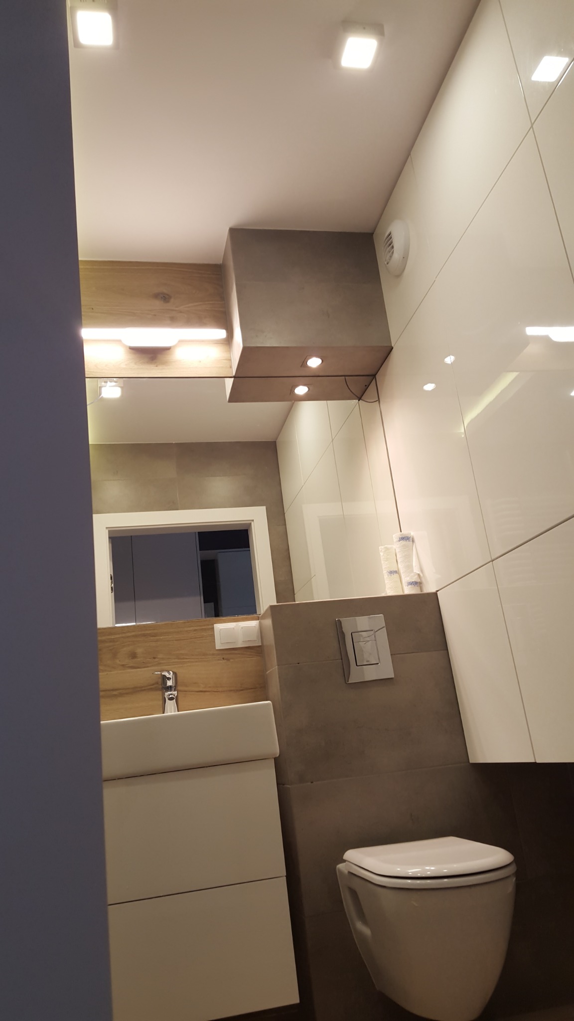 Nowoczesna łazienka z podwieszaną toaletą, umywalką na szafce i lustrem z oświetleniem LED, wykończona białymi i szarymi płytkami ceramicznymi.