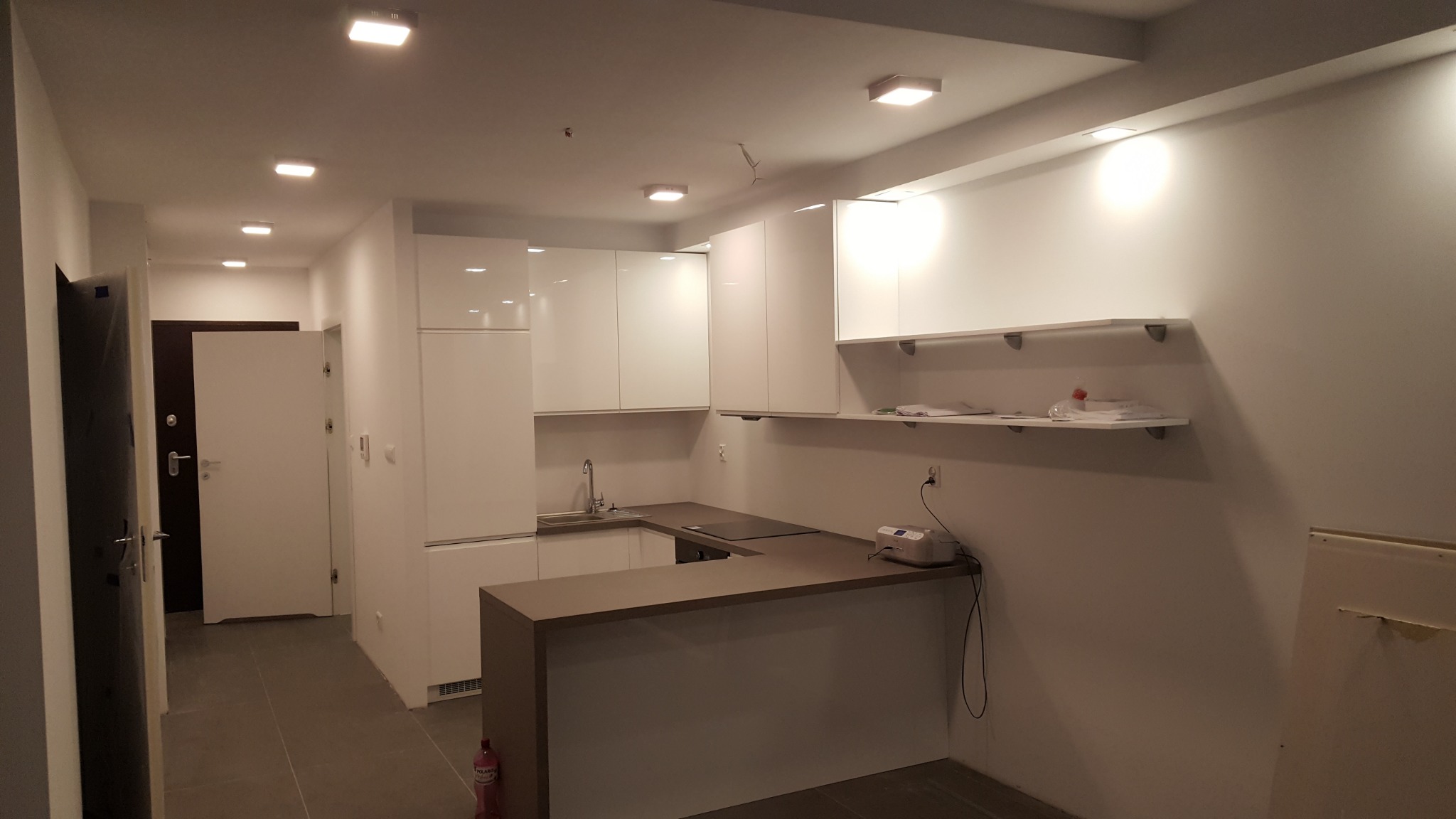 Nowoczesna kuchnia z białymi, lakierowanymi szafkami, brązowym blatem i otwartymi półkami, oświetlona punktowymi lampami, widok na drzwi wejściowe w tle.