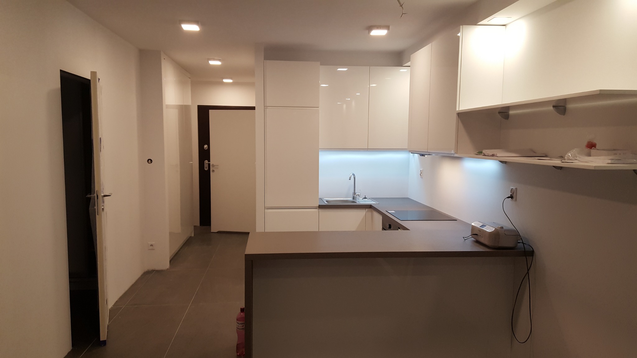 Jasna kuchnia z białymi szafkami i szarobrązowym blatem, podświetleniem LED pod szafkami wiszącymi, widok na otwarte drzwi wejściowe i butelkę wody mineralnej przy podłodze.