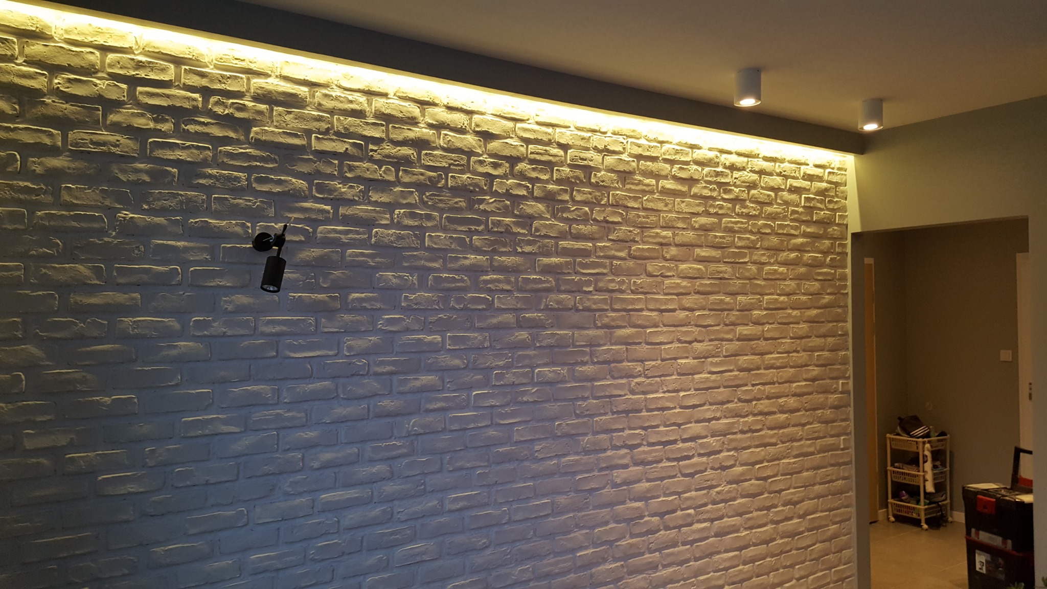 Ściana z białej cegły dekoracyjnej, oświetlona od góry listwą LED, z zamontowanym punktowym reflektorem na ścianie, widoczne również dwa reflektory sufitowe.