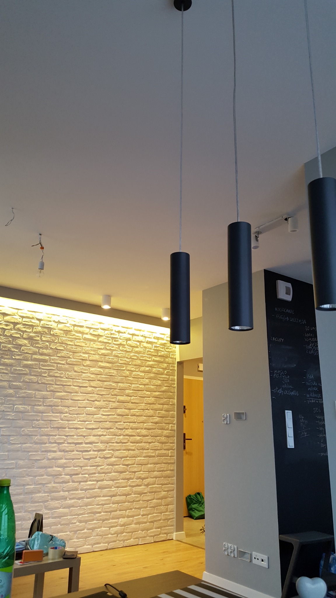 Trzy czarne cylindryczne lampy wiszące na tle białej ceglanej ściany z oświetleniem LED i tablicy kredowej z notatkami.