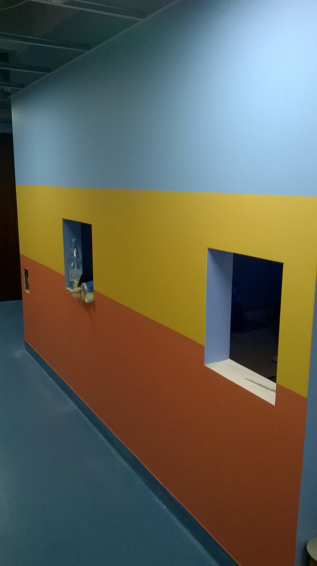 Ściana pomalowana w poziome pasy w kolorach: niebieskim, żółtym i pomarańczowym, z dwoma otworami w ścianie, z których jeden zawiera butelkę z wodą i inne przedmioty