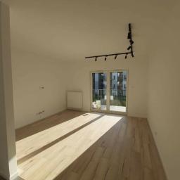 Mastbo - Puste, jasne pomieszczenie po remoncie z panelami podłogowymi imitującymi drewno, białymi ścianami, grzejnikiem i czarnym, modernistycznym oświetleniem sufitowym.