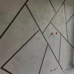 Dekoracyjna ściana z szarą, teksturowaną farbą i geometrycznymi, czarnymi liniami tworzącymi nieregularne kształty, widoczne gniazdka elektryczne.