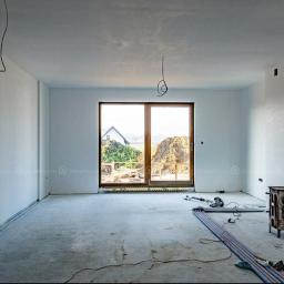 Mastbo - Surowy stan wnętrza z widocznymi przewodami elektrycznymi zwisającymi z sufitu, przygotowane do dalszych prac wykończeniowych, z oknem na ogród i stertą ziemi na zewnątrz.