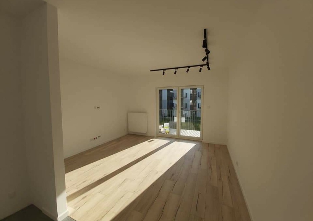 Puste, jasne pomieszczenie po remoncie z panelami podłogowymi imitującymi drewno, białymi ścianami, grzejnikiem i czarnym, modernistycznym oświetleniem sufitowym.