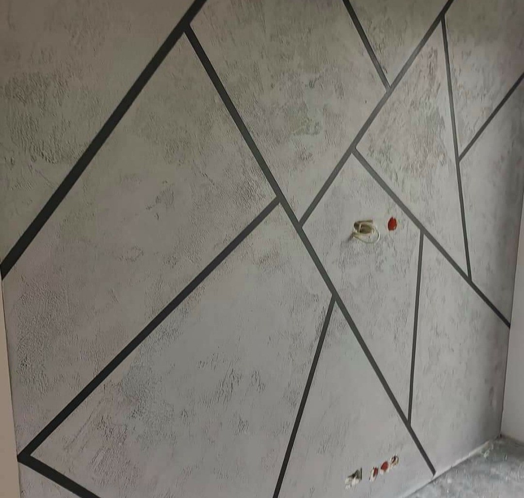 Dekoracyjna ściana z szarą, teksturowaną farbą i geometrycznymi, czarnymi liniami tworzącymi nieregularne kształty, widoczne gniazdka elektryczne.