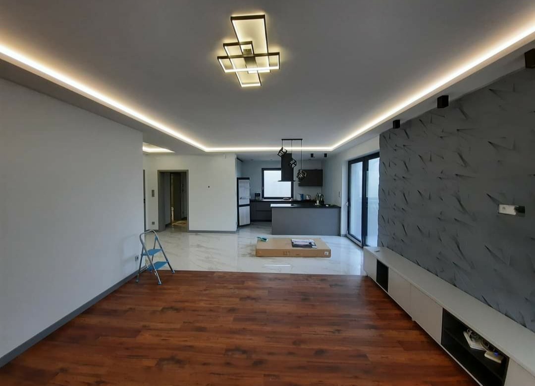 Przestronne wnętrze po remoncie z drewnianą podłogą, dekoracyjną ścianą 3D w odcieniach szarości, podświetlanym sufitem i nowoczesną lampą. W tle widoczny aneks kuchenny z szarą zabudową i wyspą.