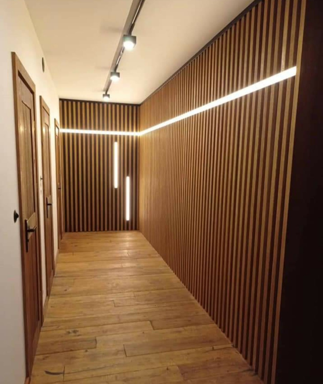 Korytarz z drewnianymi lamelami na ścianach i suficie, oświetlony liniowymi lampami LED i reflektorami na szynie, z drewnianą podłogą i drzwiami po lewej stronie.