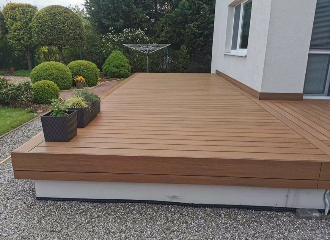 Nowoczesny taras z desek kompozytowych w kolorze jasnego drewna, z dwoma czarnymi donicami z roślinami, przylegający do białego budynku mieszkalnego otoczony zielenią.