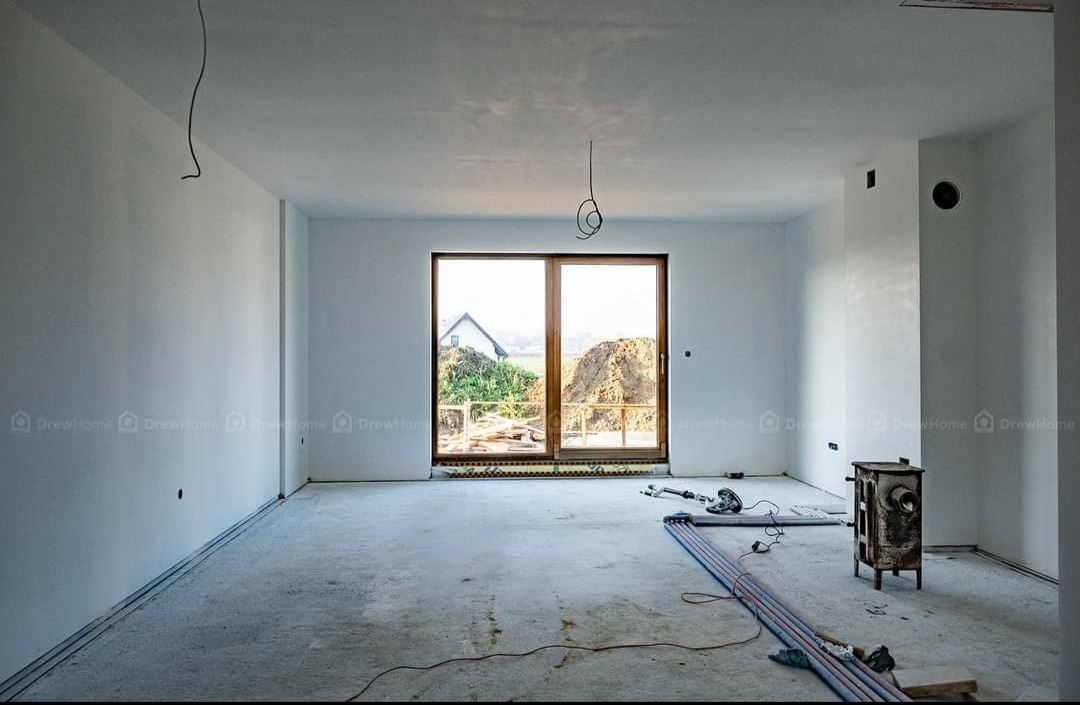 Surowy stan wnętrza z widocznymi przewodami elektrycznymi zwisającymi z sufitu, przygotowane do dalszych prac wykończeniowych, z oknem na ogród i stertą ziemi na zewnątrz.