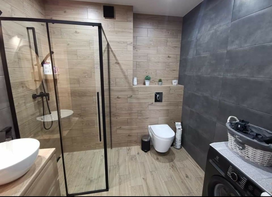 Nowoczesna łazienka z kabiną prysznicową typu walk-in z czarnymi ramami, umywalką nablatową, toaletą podwieszaną, drewnopodobnymi płytkami na ścianach i podłodze oraz szarą ścianą z dużych płyt...
