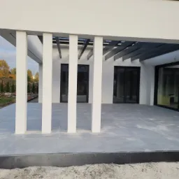 Nowoczesny taras z szarych płyt kamiennych, zadaszony pergolą z białymi słupami, widok na ogród i dom z dużymi oknami, obrzeże tarasu z czarnych kostek, piasek na obrzeżach.