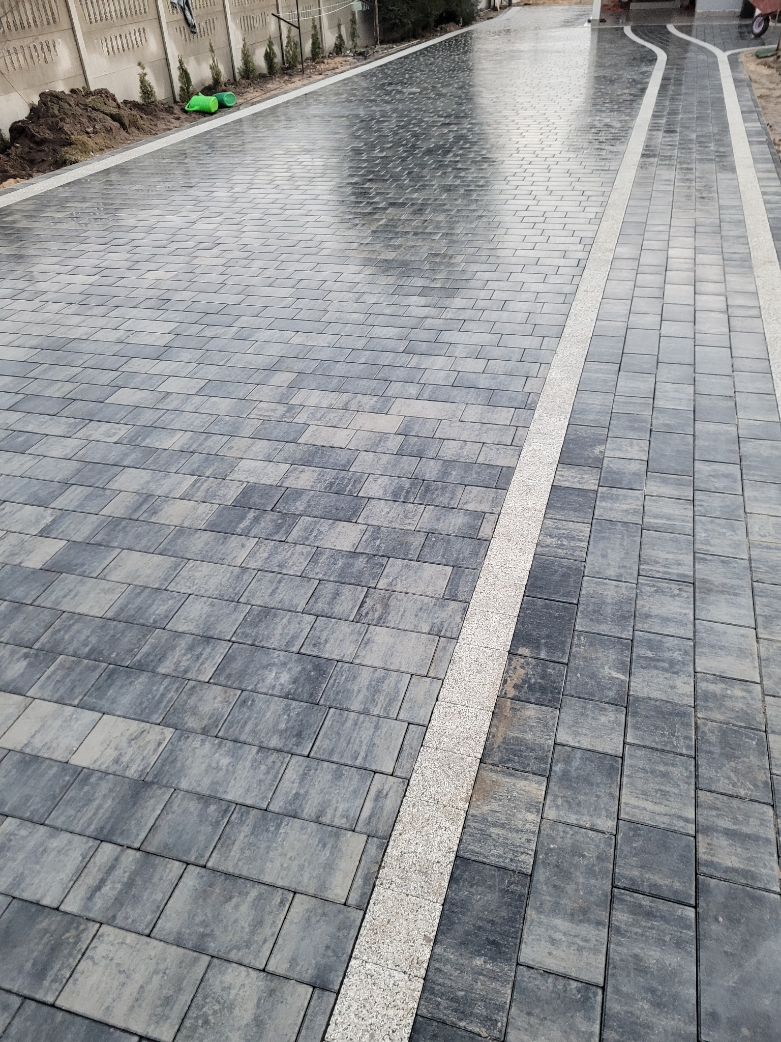 Ułożona, mokra nawierzchnia z prostokątnej kostki brukowej w odcieniach szarości, oddzielona pasem z drobnego kruszywa. Widoczny fragment betonowego ogrodzenia i roślinność w tle.