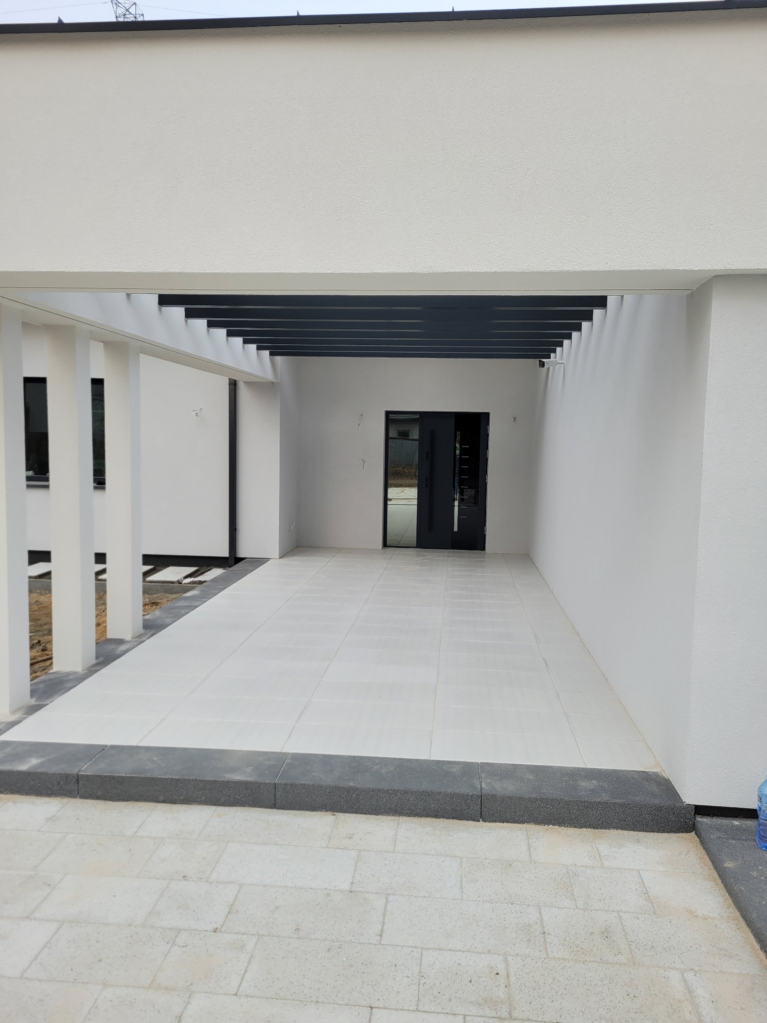Zadaszony taras z białymi płytkami i ciemnoszarą pergolą, widoczne obrzeża z kostki brukowej i wejście do budynku z ciemnymi drzwiami.