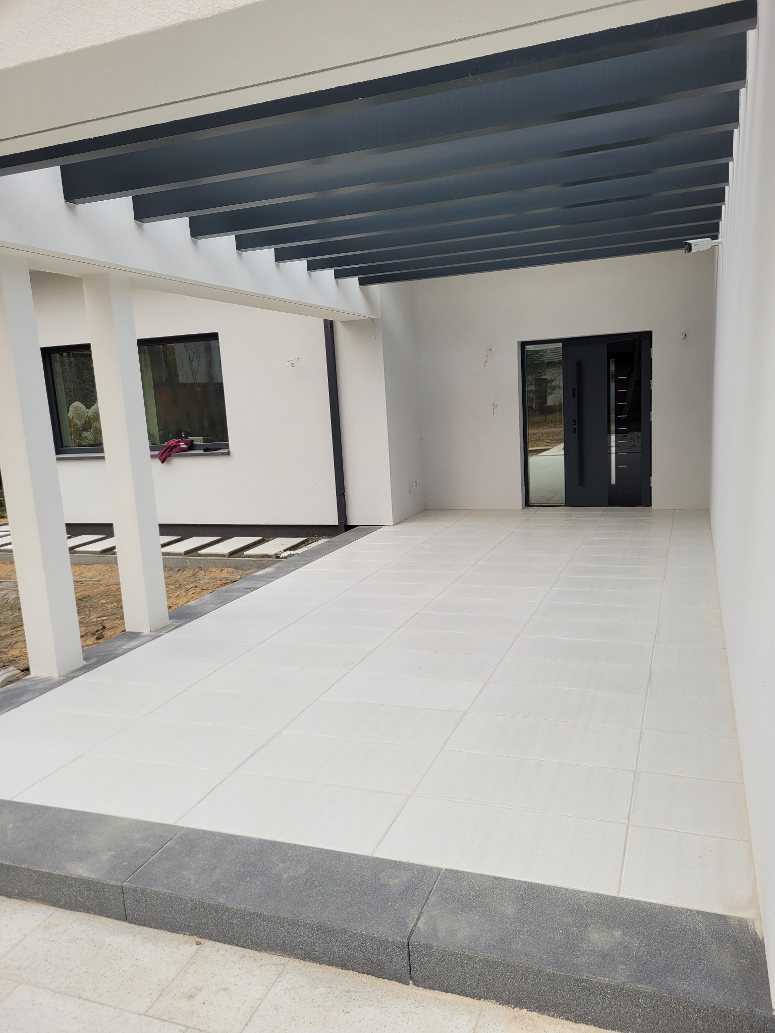 Zadaszony taras z jasnymi płytami i ciemnoszarą obwódką, widok na drzwi wejściowe i fragment ogrodu z elementami ścieżki.