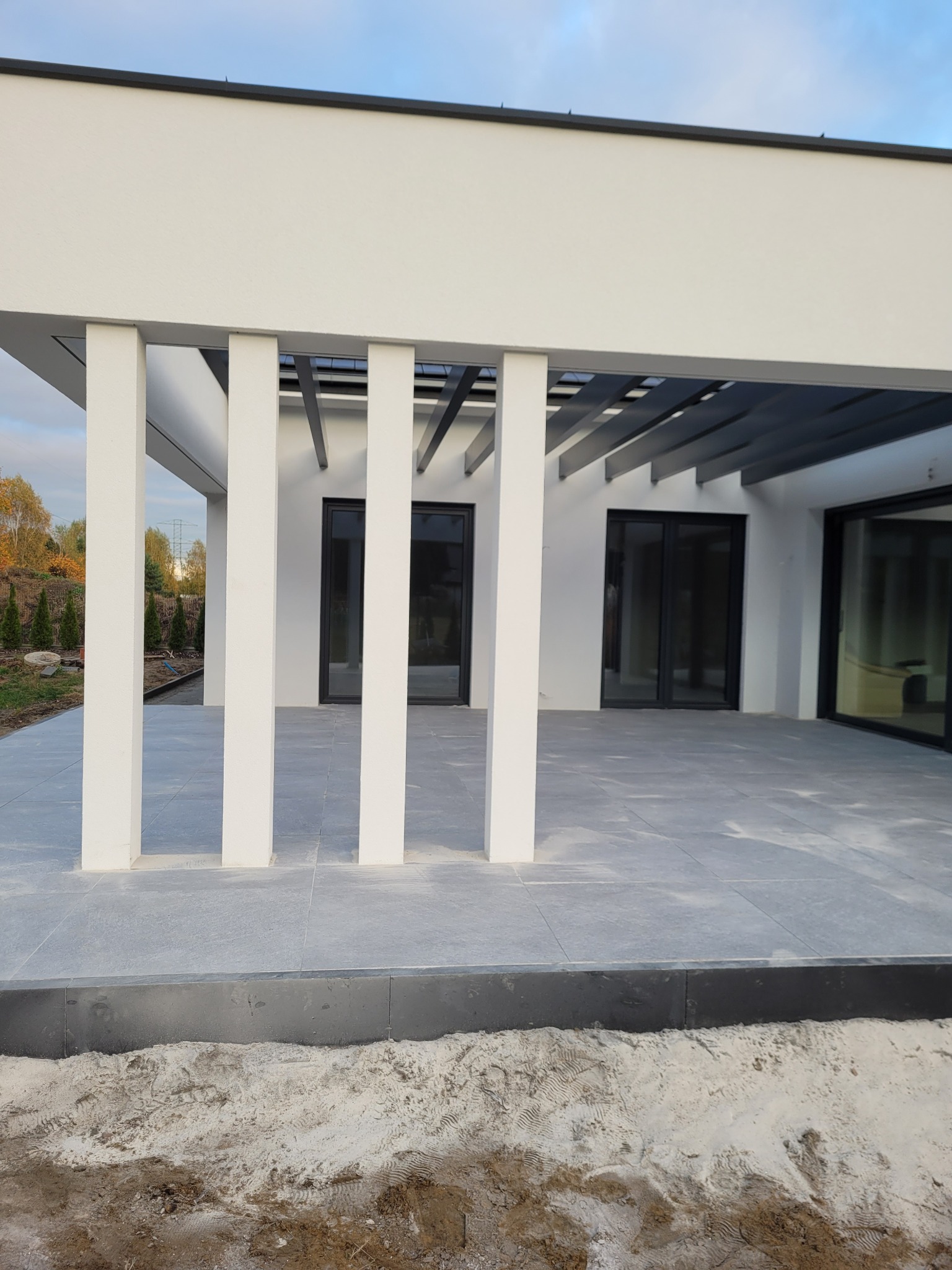 Nowoczesny taras z szarych płyt kamiennych, zadaszony pergolą z białymi słupami, widok na ogród i dom z dużymi oknami, obrzeże tarasu z czarnych kostek, piasek na obrzeżach.