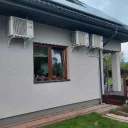 Cztery jednostki klimatyzacji Sinclair DC Inverter zamontowane na elewacji domu jednorodzinnego z brązowymi oknami, widoczne panele słoneczne na dachu i fragment tarasu.