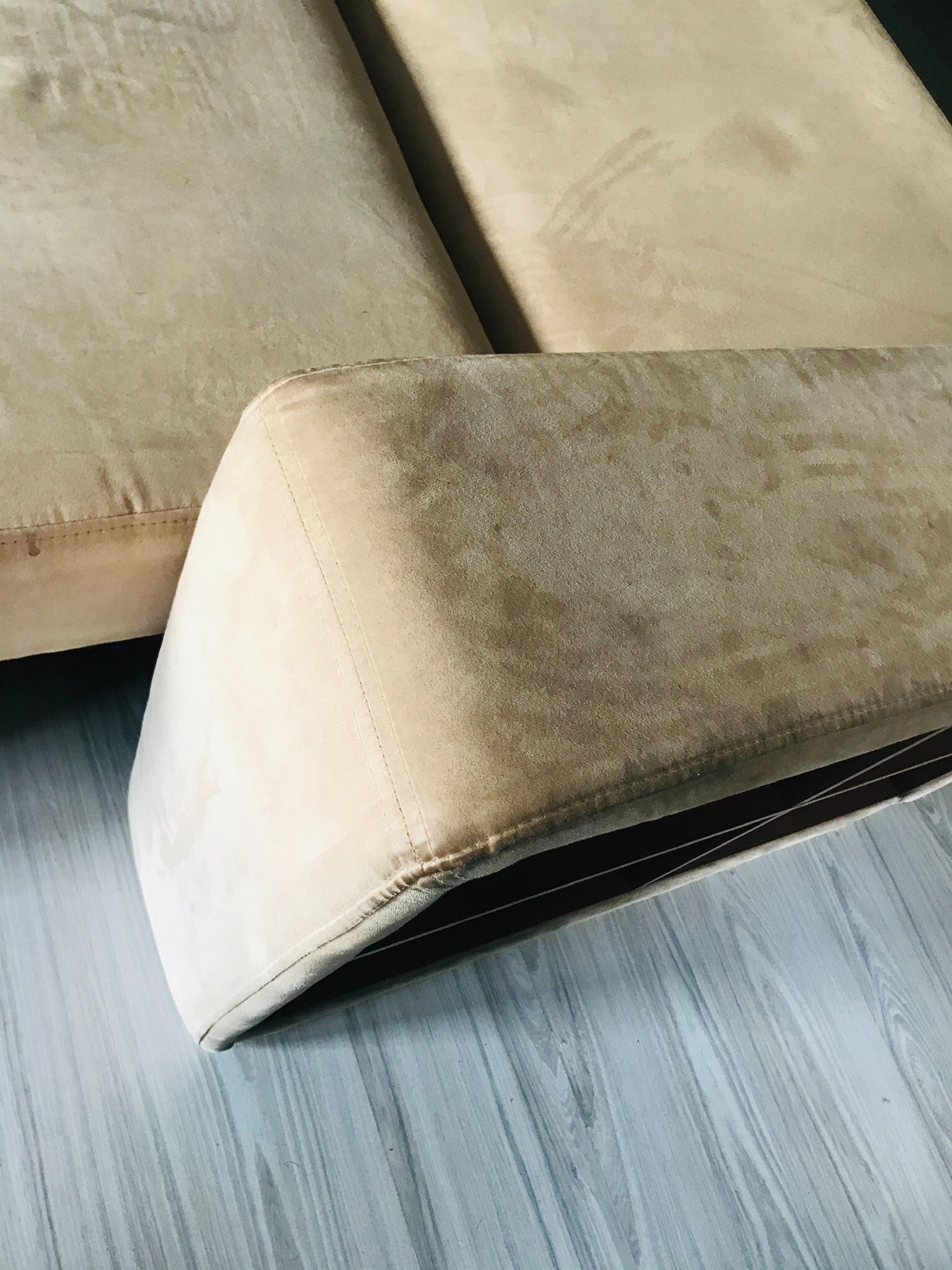 Fragment beżowej, welurowej kanapy z widocznym szwem i jasną podłogą imitującą drewno.