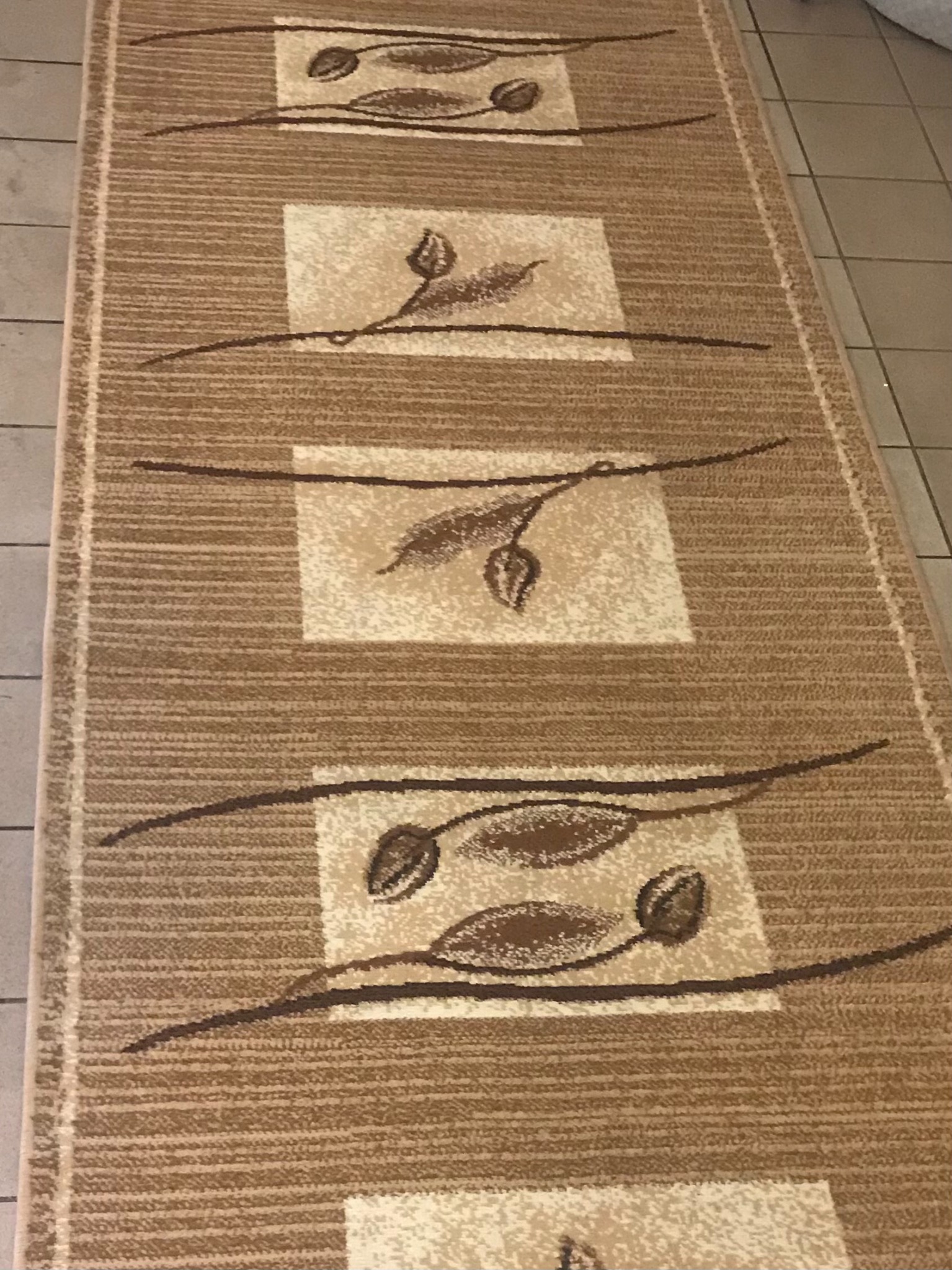 Beżowy dywanik z geometrycznym wzorem liści na jasnych kwadratach, ułożony na kafelkach.