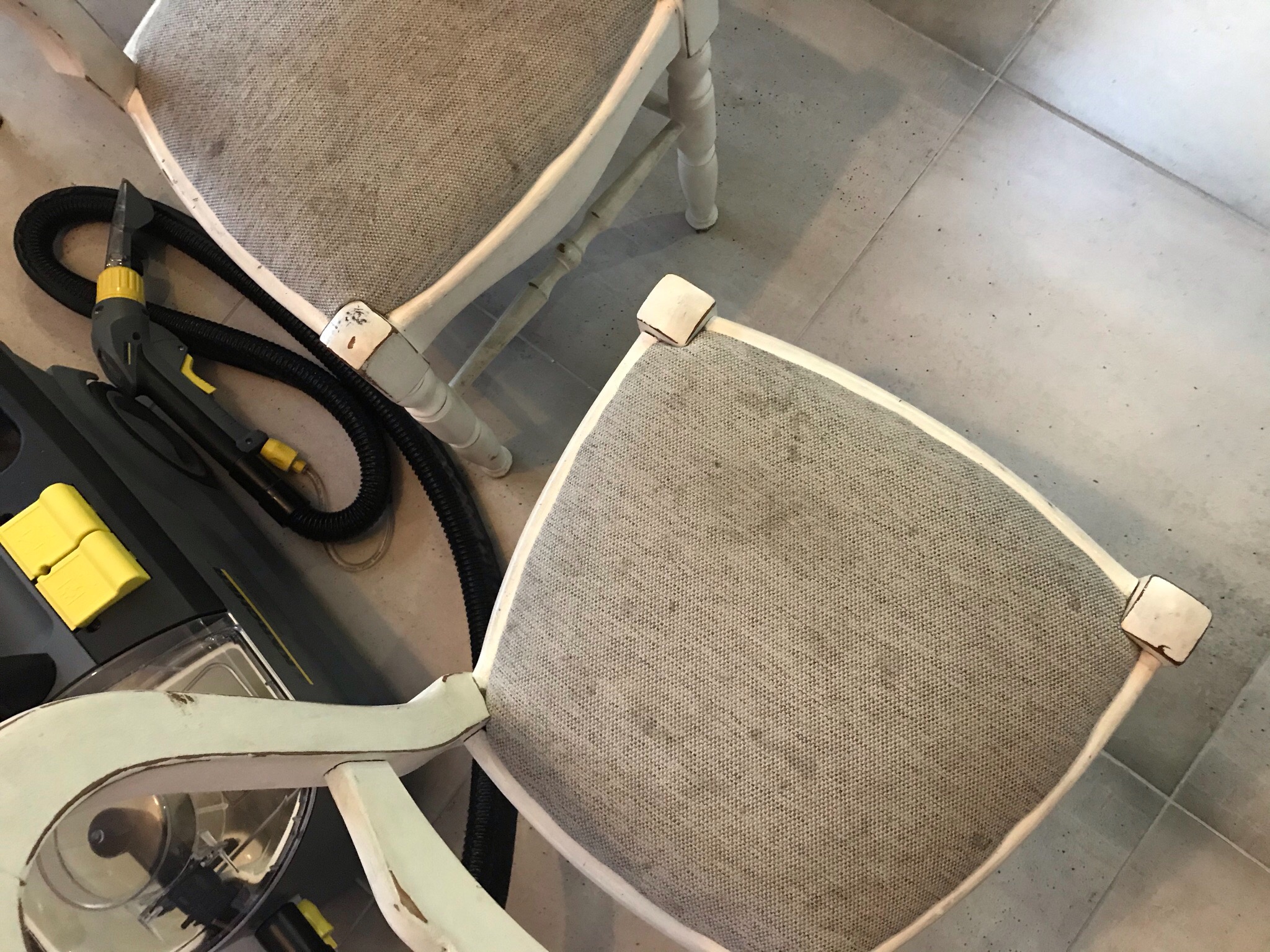 Dwa krzesła z tapicerowanymi siedziskami przed czyszczeniem, widoczny odkurzacz piorący z dyszą ssącą, zabrudzenia na tkaninie.