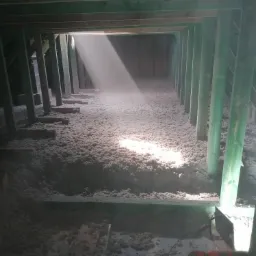 Poddasze z drewnianą konstrukcją wypełnione szarą izolacją celulozową, oświetlone smugą światła wpadającą z góry.