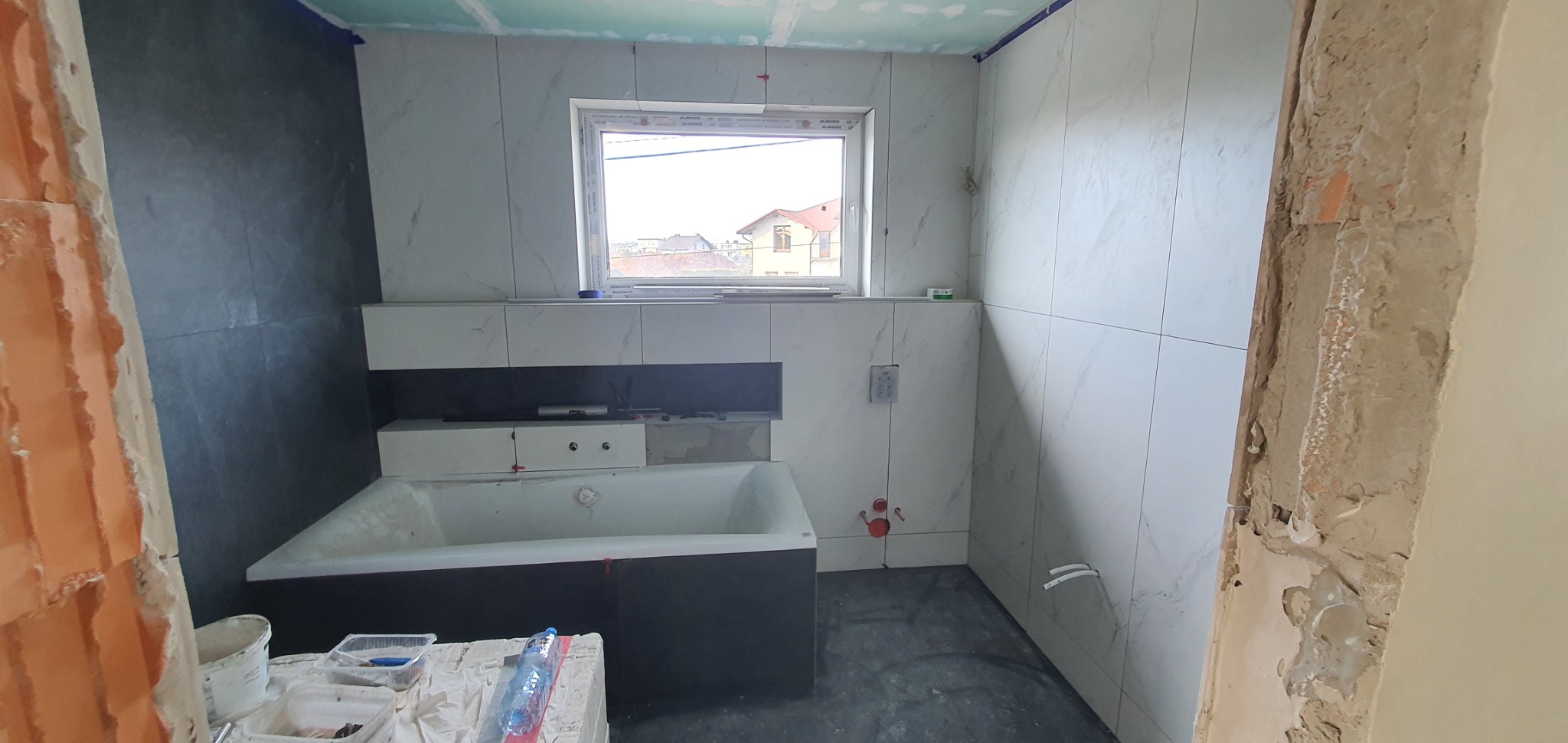 Wnętrze łazienki w trakcie prac wykończeniowych, widoczna wanna obudowana czarną płytą, białe płytki imitujące marmur na ścianach, okno z widokiem na zabudowę jednorodzinną, fragment ściany...