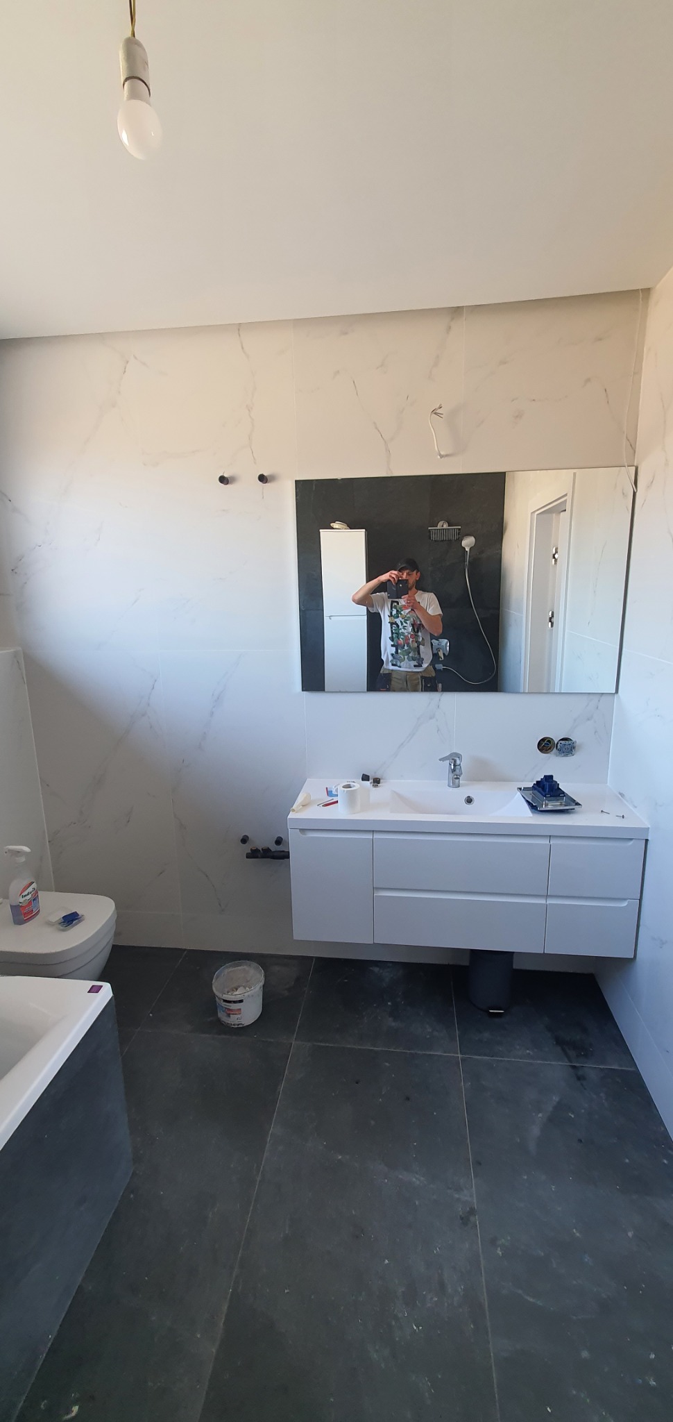 Nowoczesna łazienka w trakcie wykańczania: białe płytki imitujące marmur, szara podłoga z płytek, biała szafka pod umywalką z baterią, lustro z odbiciem osoby robiącej zdjęcie, widoczne narzędzia...