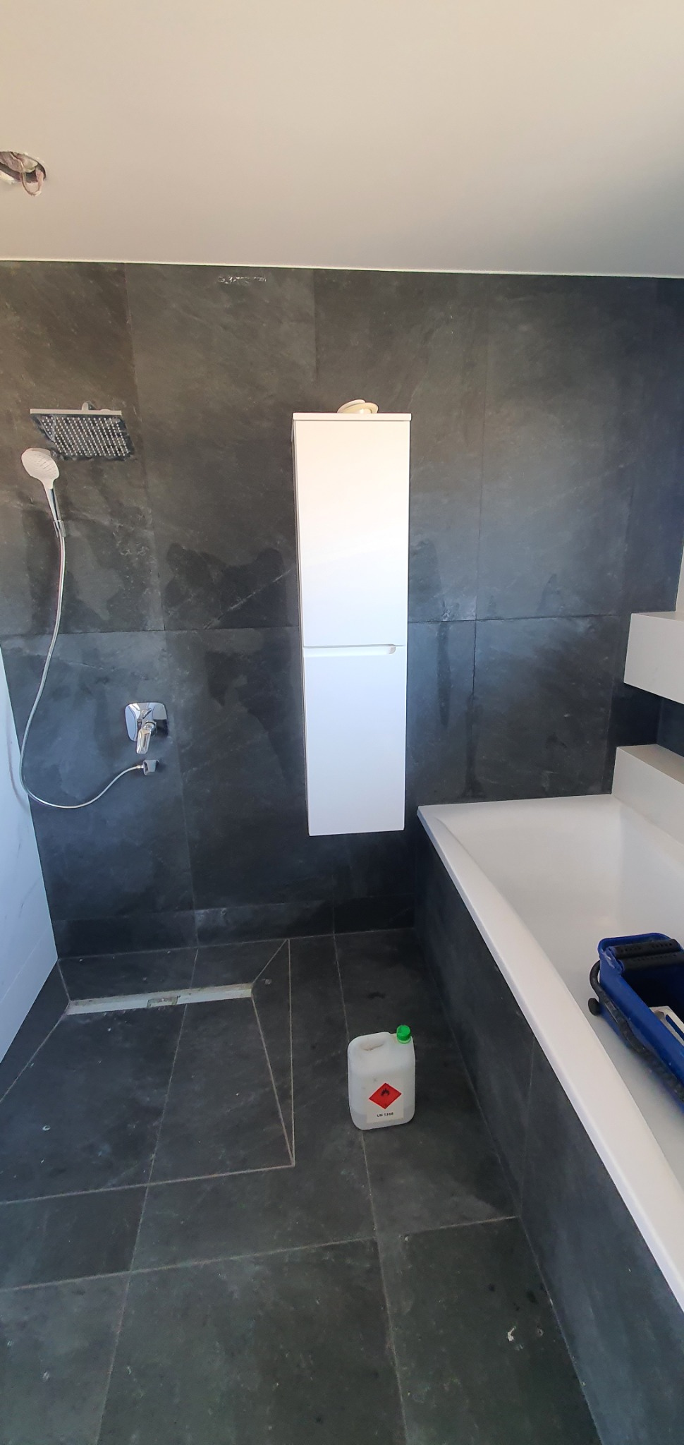 Nowoczesna łazienka z ciemnymi, marmurkowymi płytkami, białą szafką wiszącą, deszczownicą i pojemnikiem z płynem czyszczącym na podłodze, ukazująca etap prac wykończeniowych.
