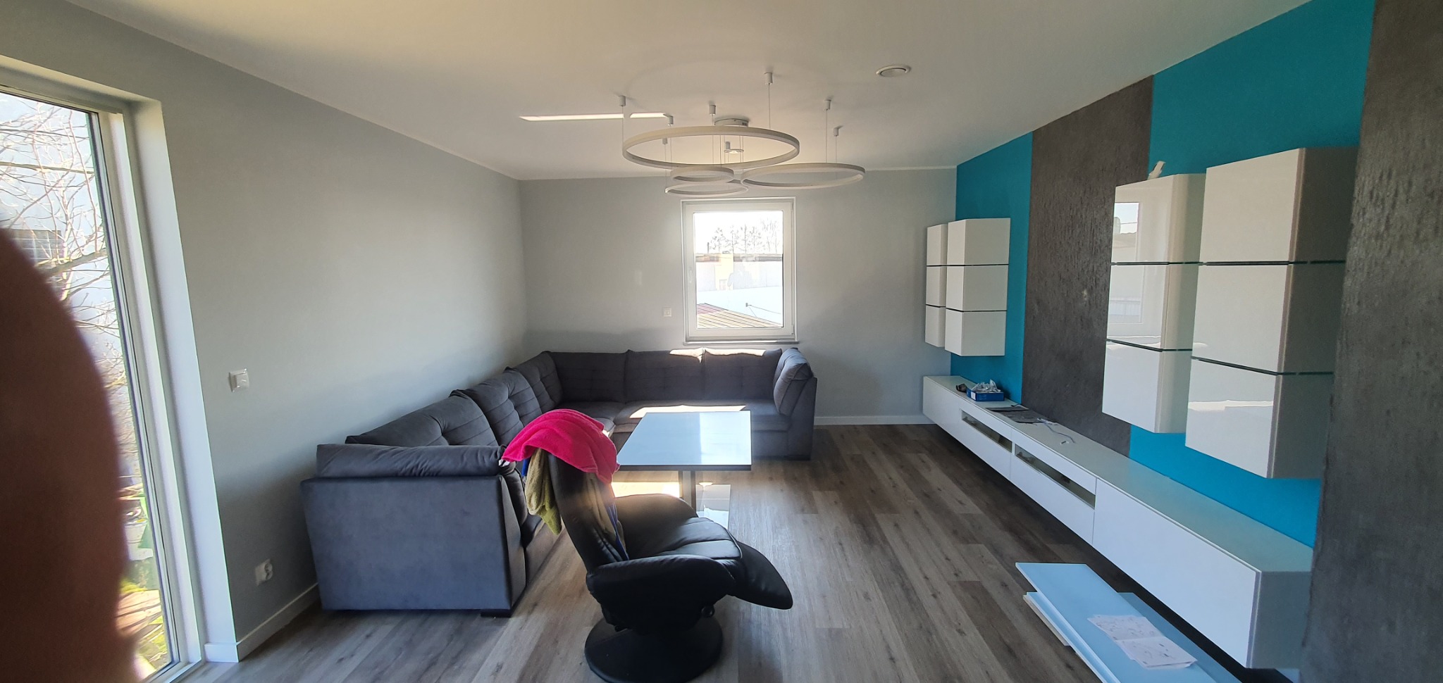 Nowoczesny salon po remoncie: szary narożnik, białe meble na tle turkusowo-szarej ściany akcentującej, parkiet w odcieniach drewna, okrągła lampa sufitowa.