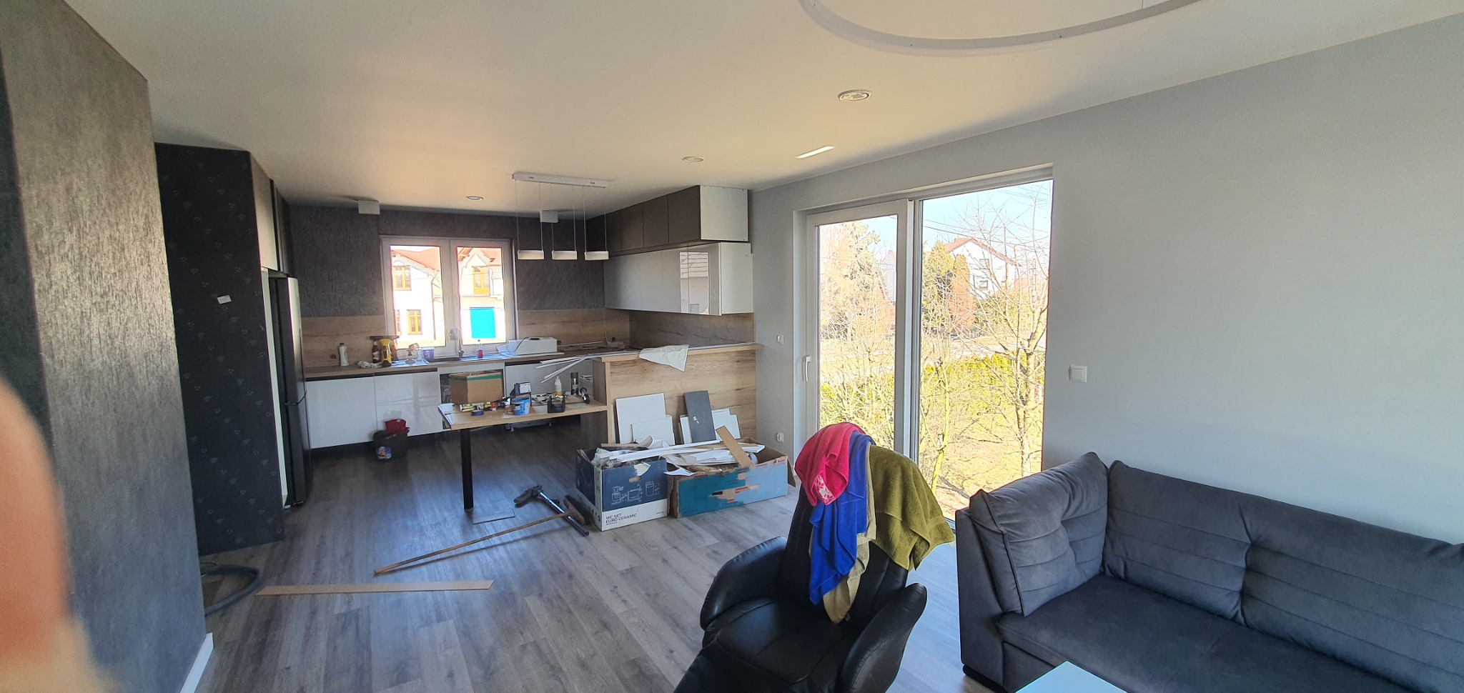 Wnętrze w trakcie prac wykończeniowych: widoczna kuchnia z zabudową, narzędzia i materiały budowlane na stole, szare panele podłogowe, sofa i fotel z kolorowymi ręcznikami w tle okna z widokiem...