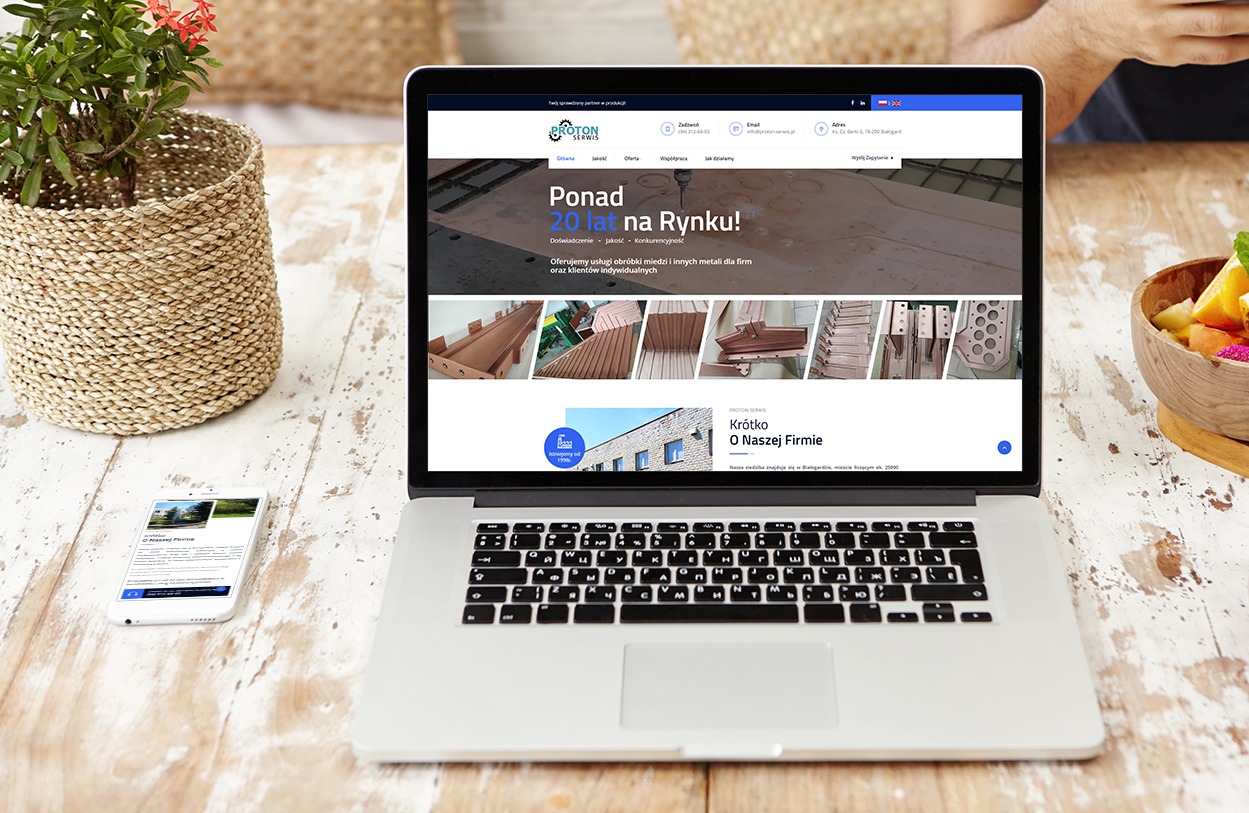 Laptop wyświetlający stronę internetową firmy Proton Serwis z ofertą obróbki metali, widoczny smartfon z podobną stroną, miseczka z owocami i roślina doniczkowa na drewnianym stole.
