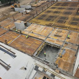 ARKBUD - Zbrojenie stropu budynku w trakcie budowy, widok z góry. Żółte szalunki, stalowe pręty zbrojeniowe, pracownicy w kaskach. Na drugim planie koparka i betonowe ściany.