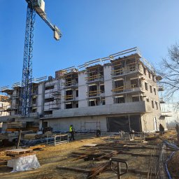 ARKBUD - Wznoszenie trzypiętrowego budynku z białego bloczka, widoczny żuraw budowlany i pracownicy w kamizelkach odblaskowych. Ujęcie z dołu, w tle błękitne niebo.