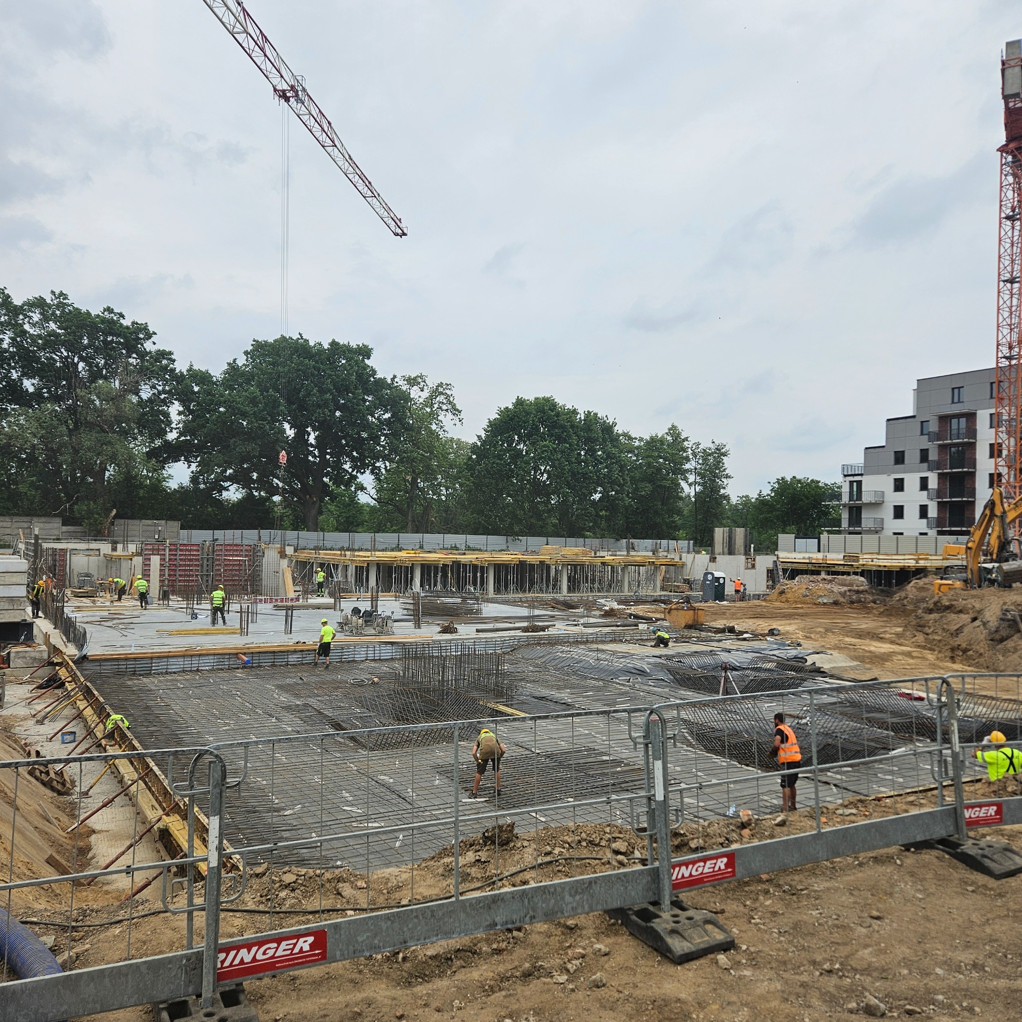 Rozległy plac budowy z widoczną konstrukcją zbrojeniową, szalunkami i pracownikami w odblaskowych kamizelkach. W tle budynek mieszkalny i żurawie.