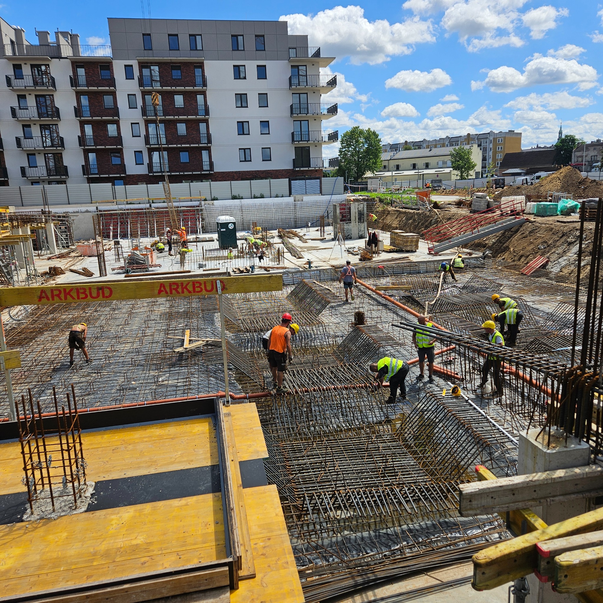 Rozległy plac budowy z widoczną siatką zbrojeniową, pracownikami w kaskach i żółtymi elementami szalunków. W tle budynek mieszkalny i błękitne niebo.