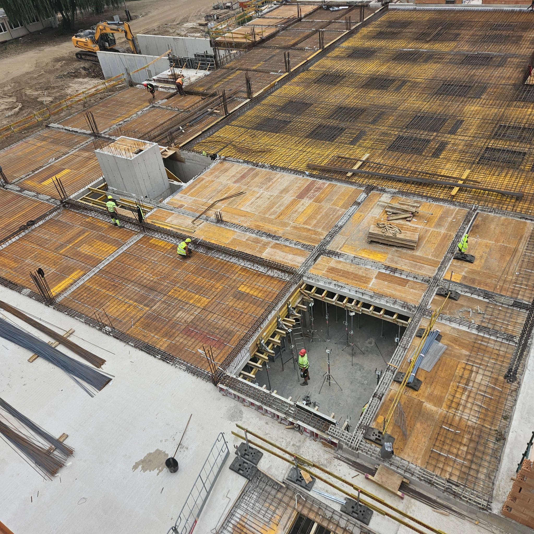 Zbrojenie stropu budynku w trakcie budowy, widok z góry. Żółte szalunki, stalowe pręty zbrojeniowe, pracownicy w kaskach. Na drugim planie koparka i betonowe ściany.