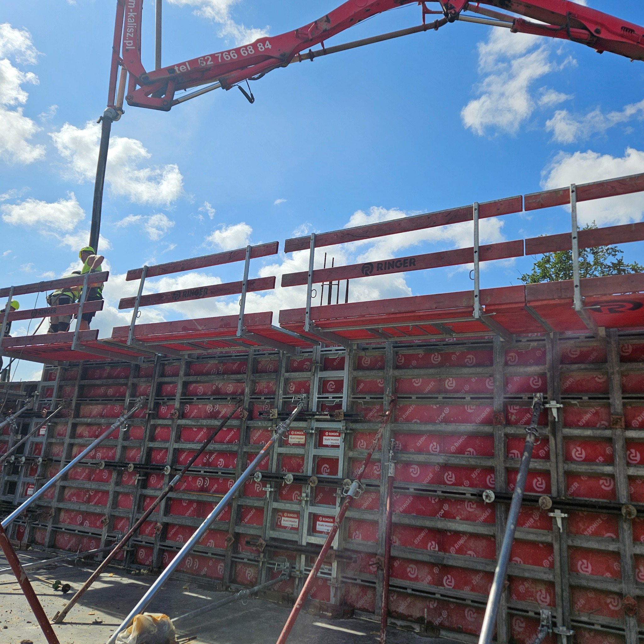 Betonowanie ściany szalunkowej z użyciem pompy do betonu. Widoczni pracownicy w kaskach. Szalunek systemowy Ringer. Błękitne niebo w tle.