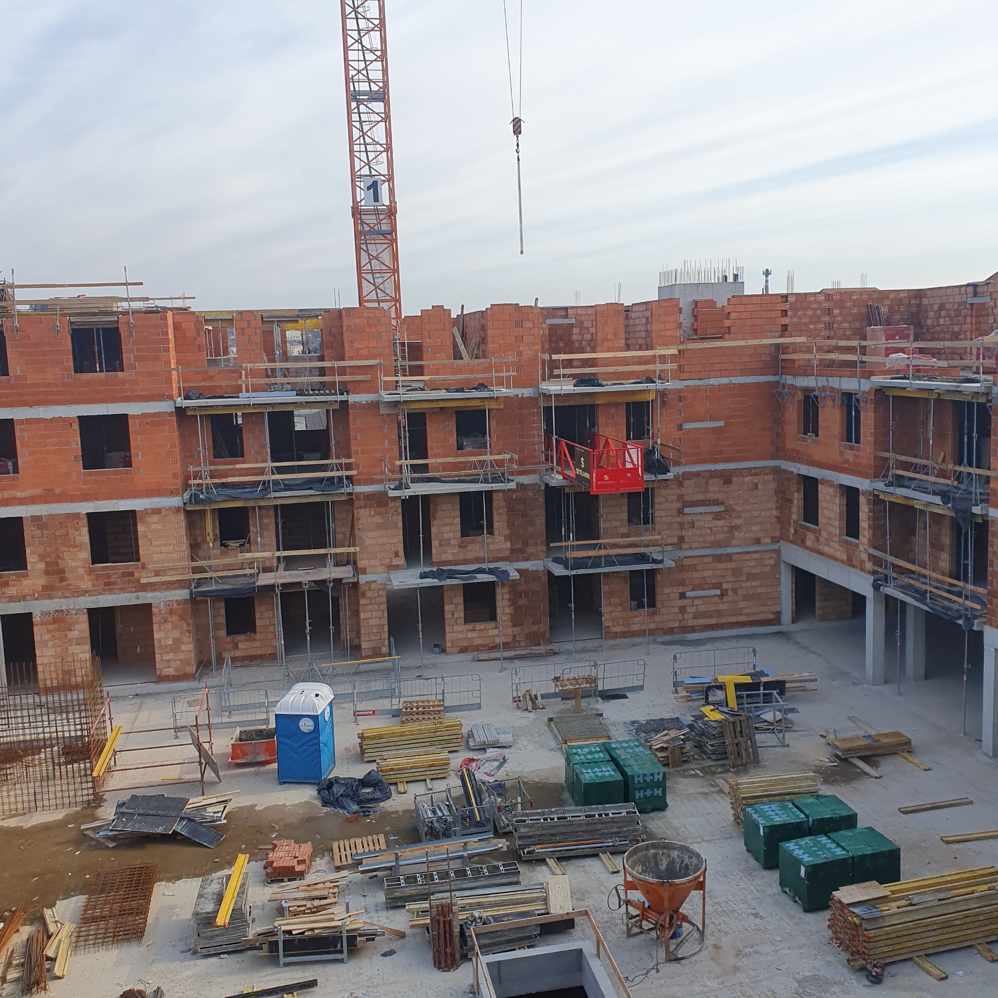 Wysoki budynek w trakcie murowania z czerwonej cegły, widok z góry. Na placu budowy widoczny dźwig, materiały budowlane i przenośna toaleta.