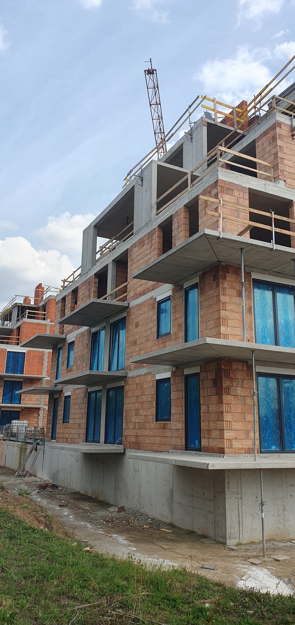 Budowa bloku mieszkalnego z czerwonej cegły, okna zabezpieczone folią, widoczny żuraw budowlany. Ujęcie z dołu, w tle pochmurne niebo.