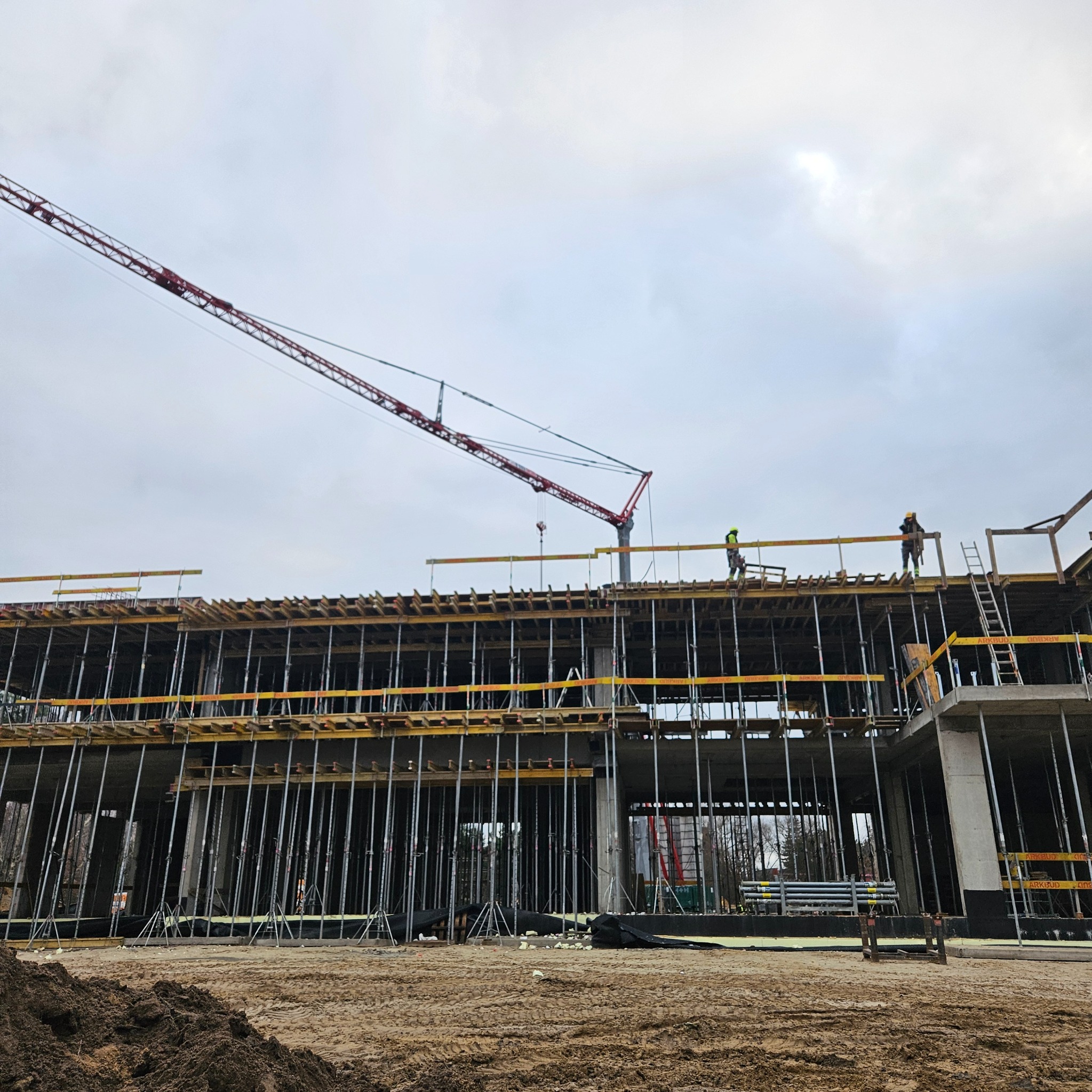 Budowa wielopiętrowego budynku w trakcie realizacji, widoczny żuraw budowlany i pracownicy na górze konstrukcji. Szalunki i podpory w dolnej części budynku.