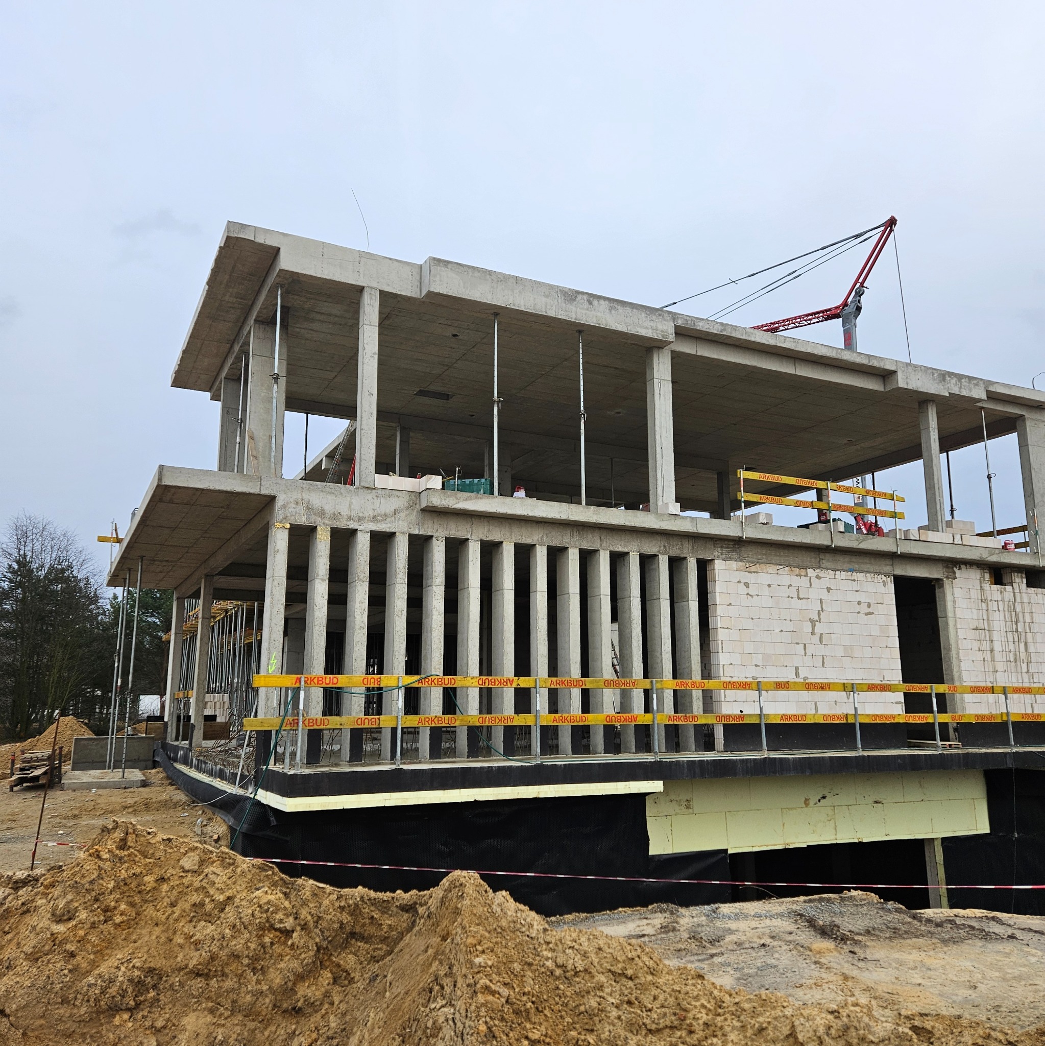 Surowy stan domu z betonu komórkowego w trakcie budowy, widoczne szalunki, podpory i żuraw na tle pochmurnego nieba. Częściowo ocieplone fundamenty.