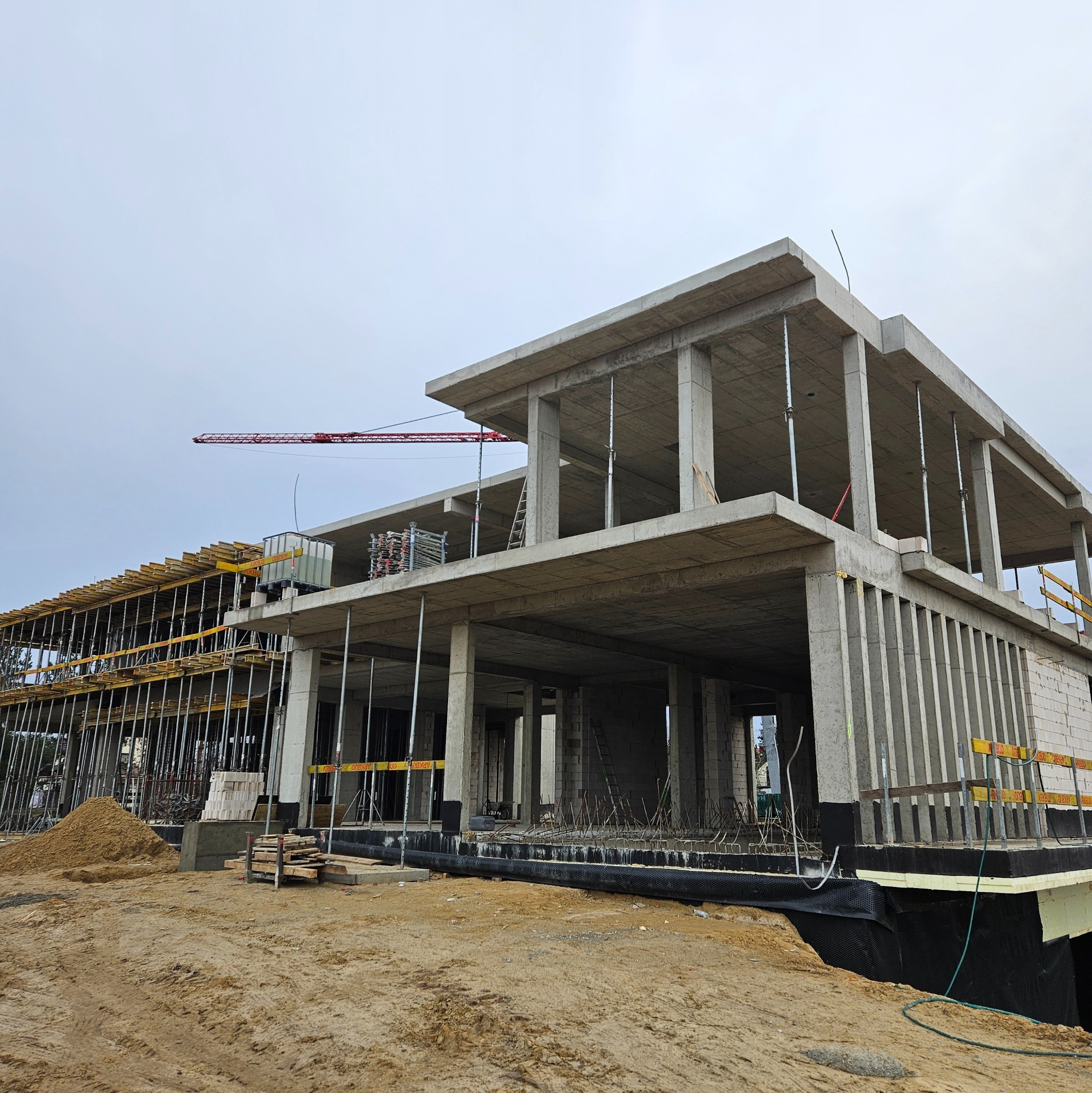 Surowy stan domu murowanego w trakcie budowy, widoczne szalunki, zbrojenia i pustaki. Na drugim planie żuraw budowlany.