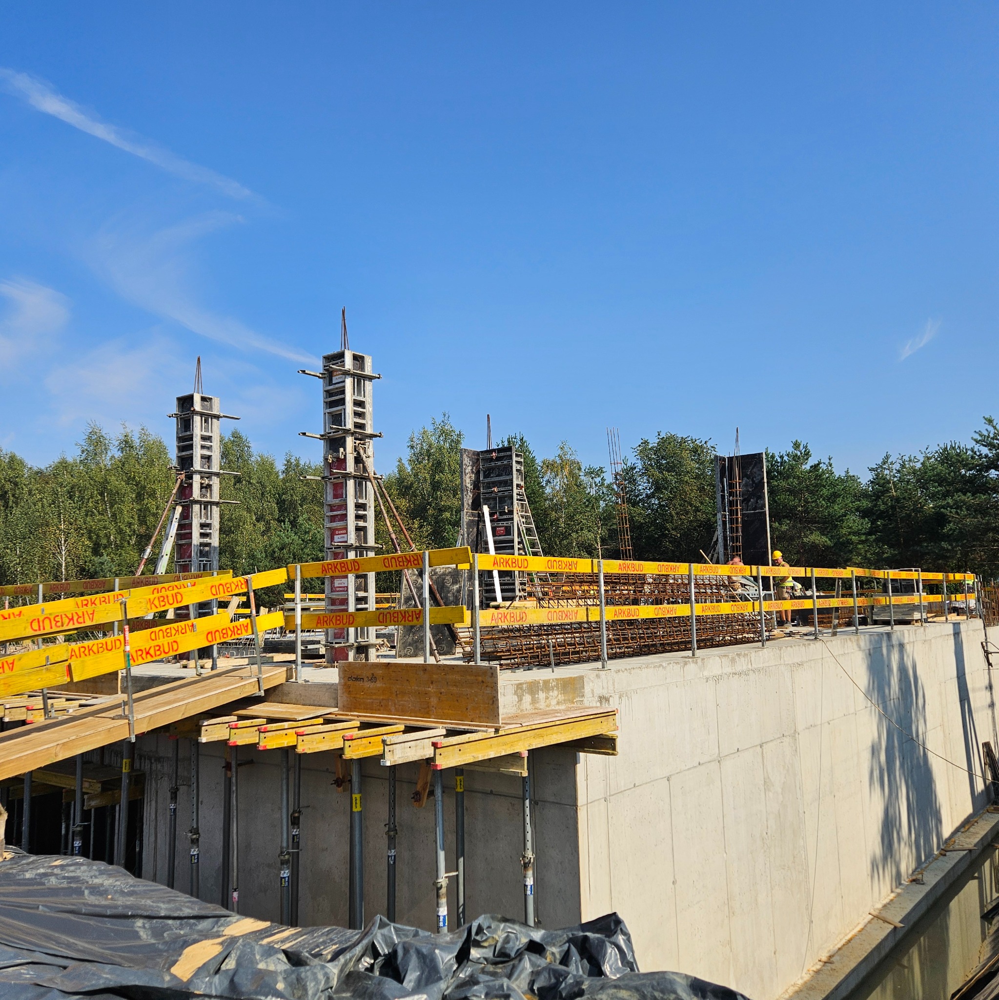 Budowa betonowej konstrukcji z żółtymi barierkami ARKBUD, widoczne zbrojenia i szalunki kolumn. W tle drzewa i błękitne niebo.