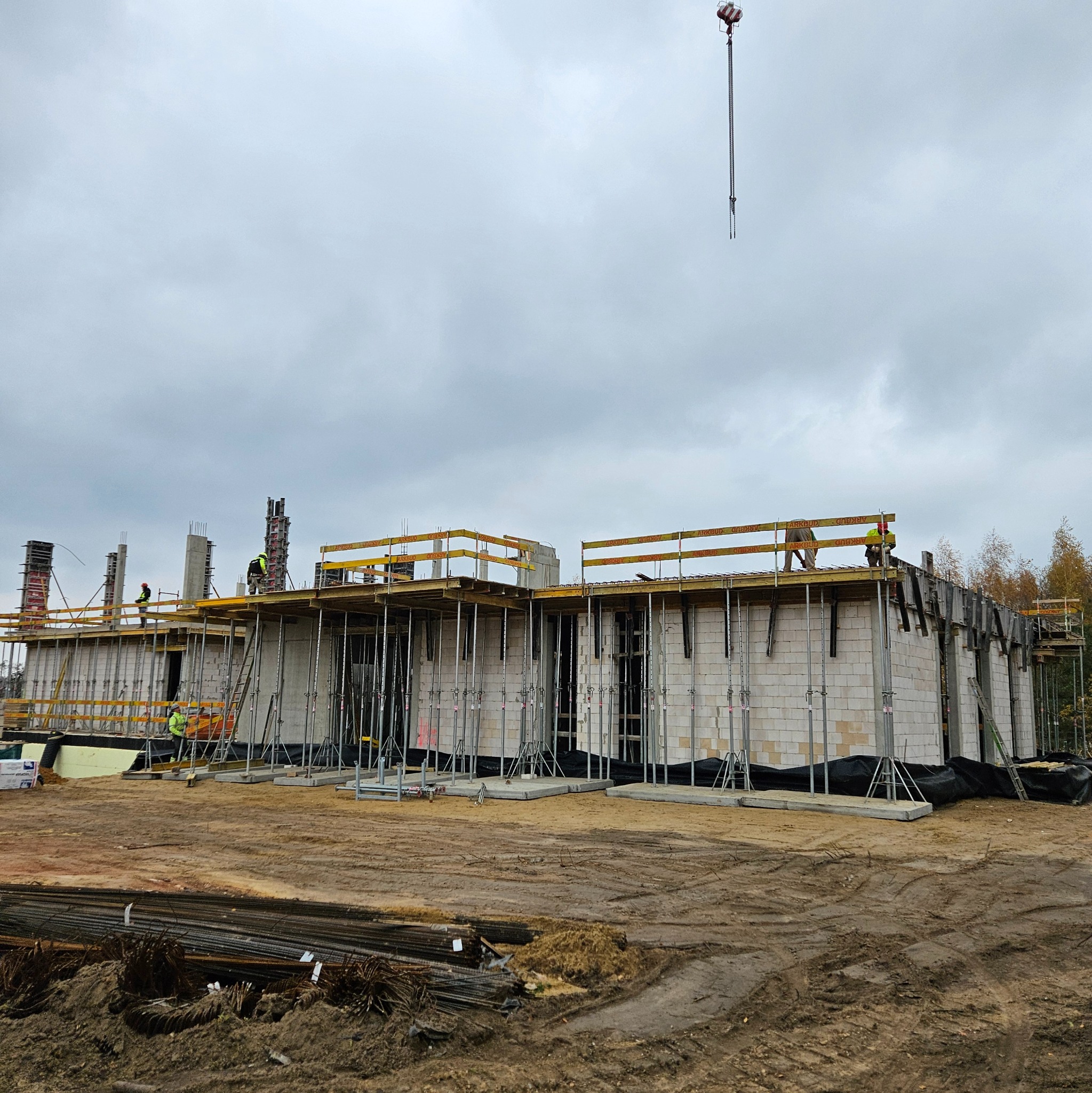 Budowa domu z białych bloczków, widoczne rusztowania, podpory i pracownicy w kaskach. Na pierwszym planie leżą pręty zbrojeniowe. W tle pochmurne niebo.