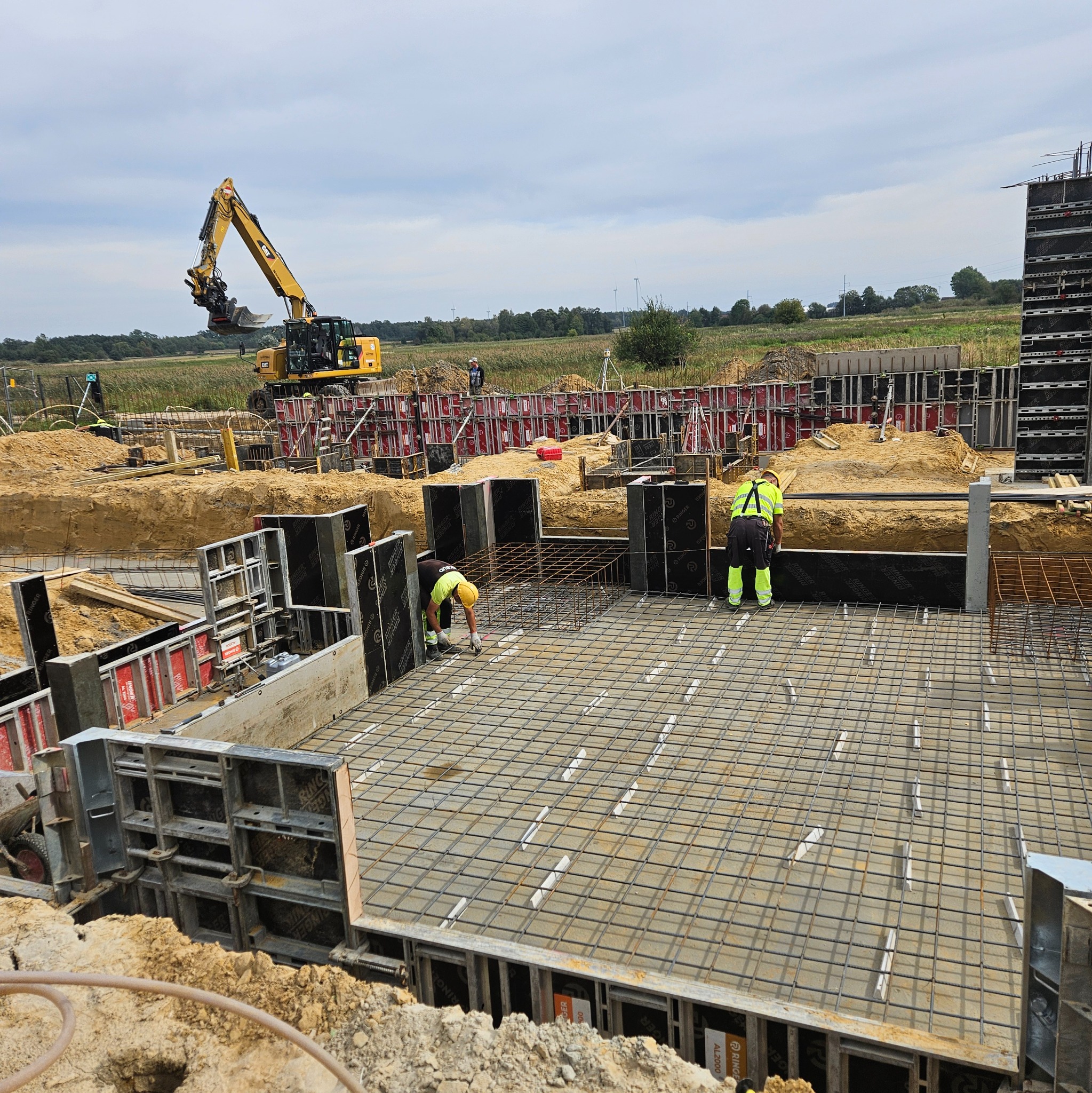 Zbrojenie fundamentów pod budowę domu jednorodzinnego. Widoczni pracownicy w odblaskowych ubraniach oraz koparka. Szalunki ustawione, teren budowy przygotowany.